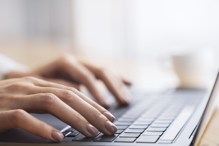 Against a blurred office scene, a close-up shot reveals a woman's hands typing on a modern laptop keyboard